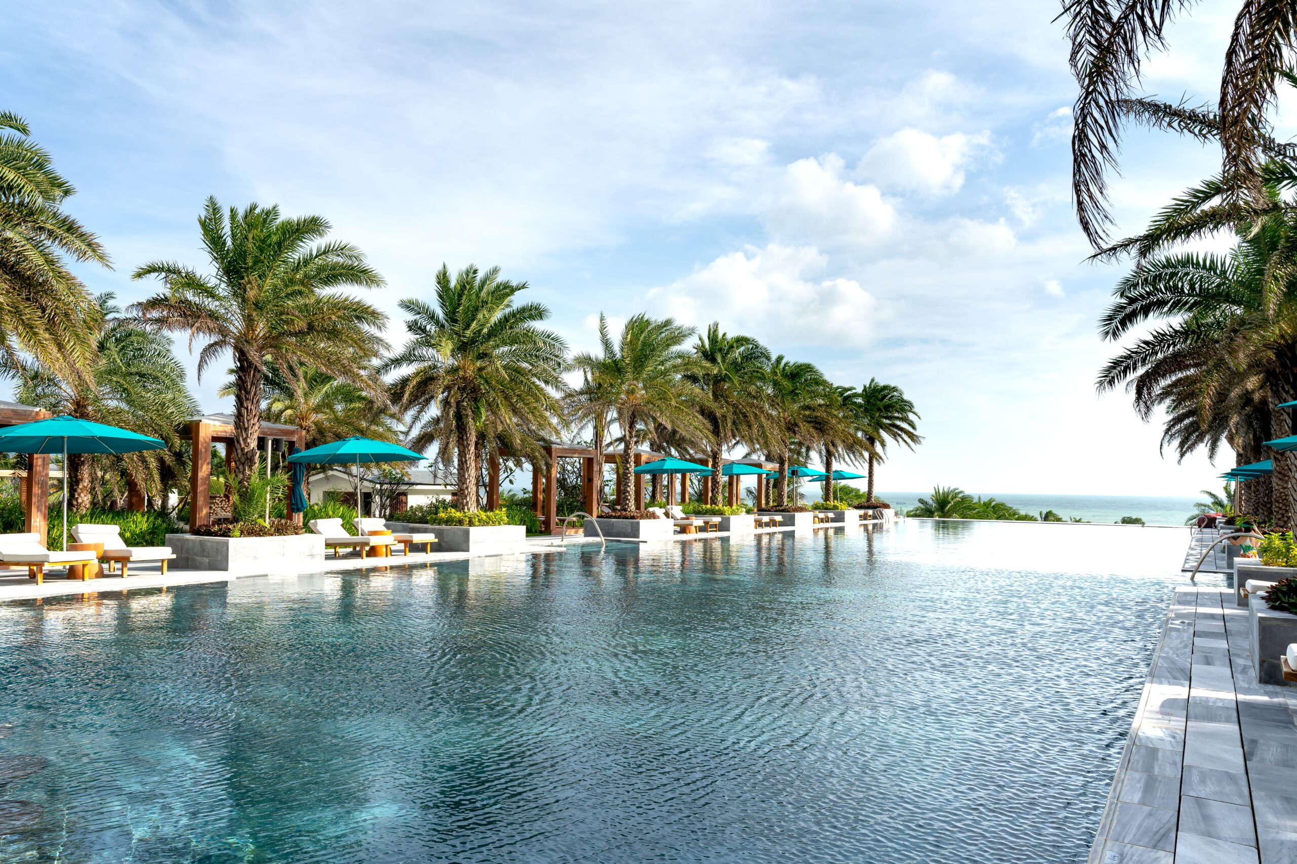 Luxury tropical resort with an infinity pool overlooking the ocean, surrounded by palm trees and elegant lounge cabanas under a bright blue sky.