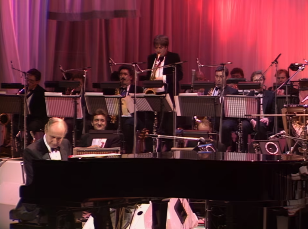 Henry Mancini performing live at the piano with his orchestra during a classic television concert featuring elegant brass, woodwinds, percussion, and lush orchestral arrangements.